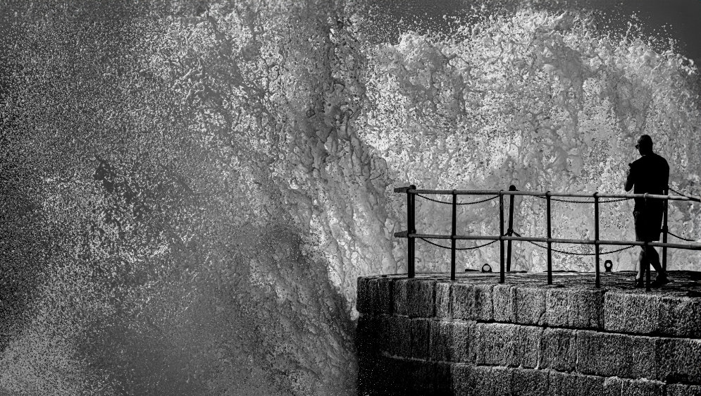 A big wave crashing against a pier, with a lone man standing at the end.
